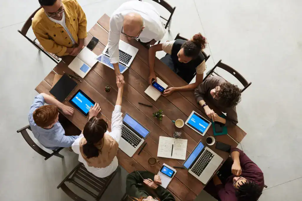Menschen bei Meeting, Handshake, Laptops und Tablets auf Tisch.