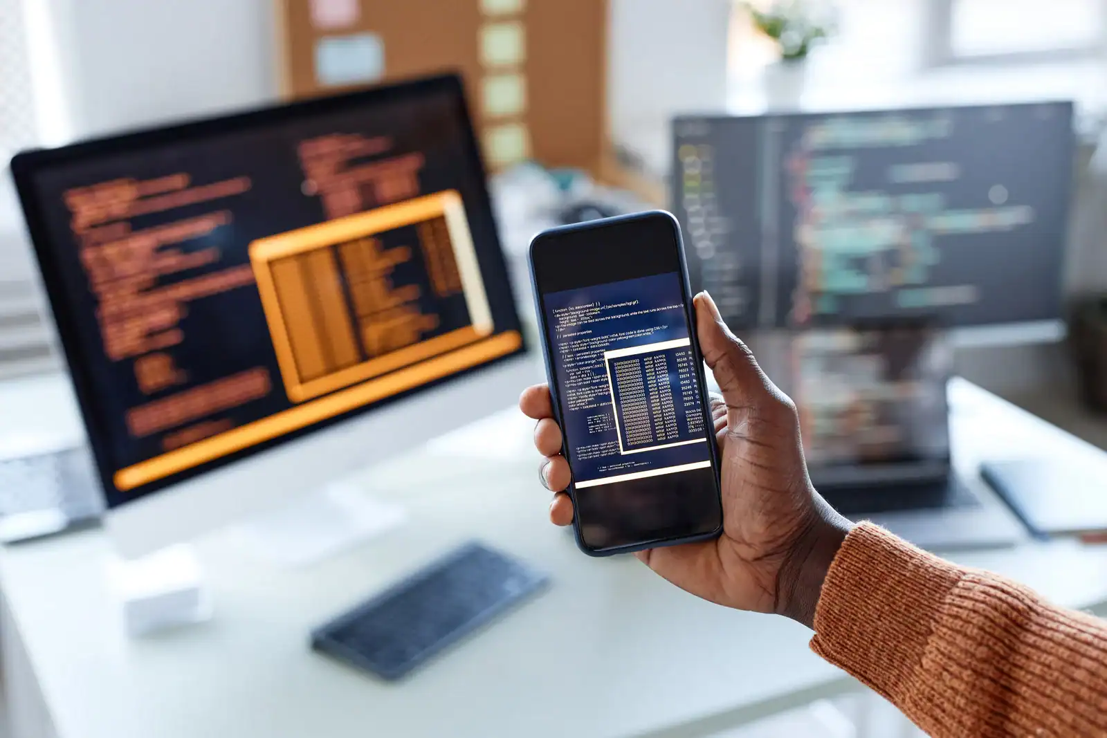 Person hält Smartphone mit Code-Editor, im Hintergrund zwei Monitore mit Programmcode.