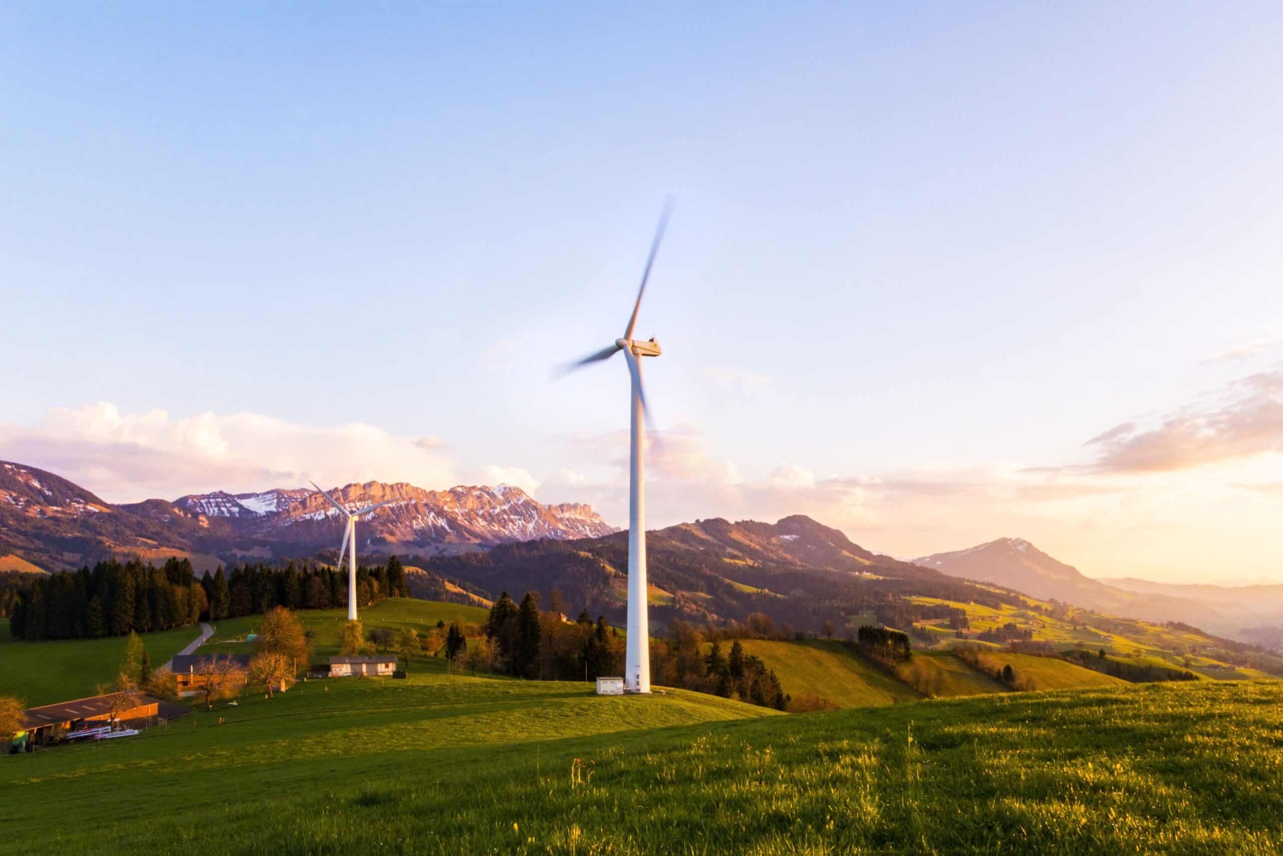 Zwei Windräder auf grünen Hügeln vor Bergen bei Sonnenuntergang.