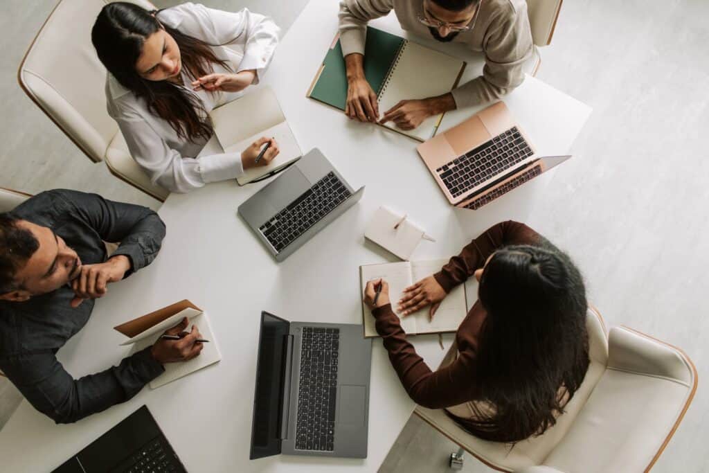 Vier Personen sitzen um einen Tisch mit Laptops, Notizbüchern und einem Tablet.